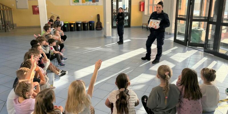 Policjantki prowadzą zajęcia profilaktyczne o bezpieczeństwie podczas ferii zimowych – dzieci siedzą na podłodze w szkolnym holu i aktywnie uczestniczą w spotkaniu.