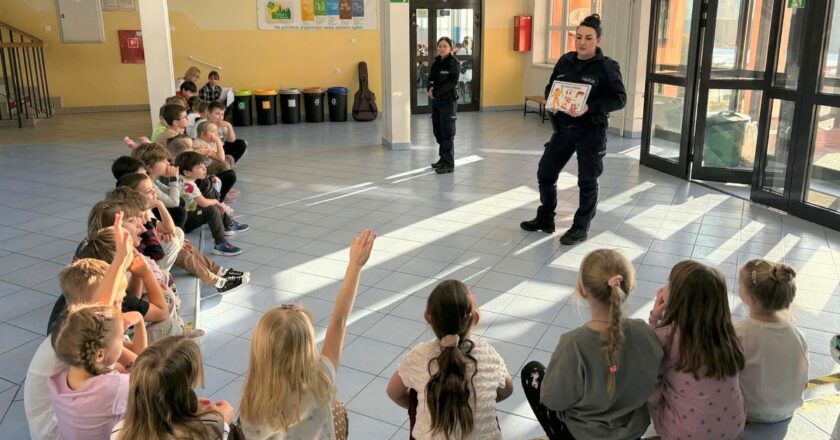 Policjantki prowadzą zajęcia profilaktyczne o bezpieczeństwie podczas ferii zimowych – dzieci siedzą na podłodze w szkolnym holu i aktywnie uczestniczą w spotkaniu.