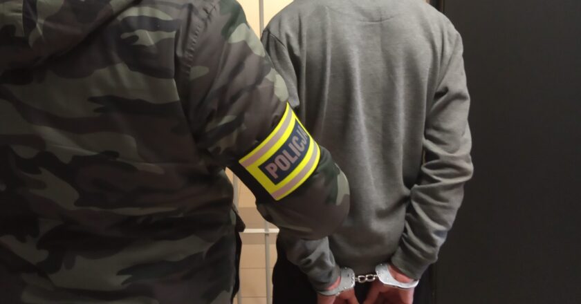 Policjant pionu kryminalnego Komendy Powiatowej Policji w Oświęcimiu prowadzi zatrzymanego mężczyznę w kajdankach na korytarzu budynku.