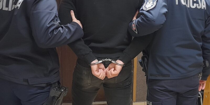 Zatrzymany mężczyzna w kajdankach, prowadzony przez dwóch policjantów w umundurowaniu, widok od tyłu w pomieszczeniu komendy Policji.