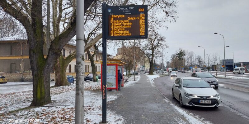 Próbnik pasywny do pomiaru dwutlenku azotu (NO₂) zamontowany przy ul. Powstańców Śląskich w Oświęcimiu, obok przystanku autobusowego i ruchliwej jezdni. W tle widoczny wyświetlacz informacji pasażerskiej „Dworzec PKP”, zabudowa miejska oraz samochody w ruchu drogowym – miejsce monitoringu zanieczyszczeń komunikacyjnych.