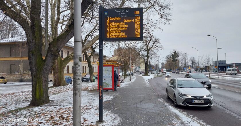Próbnik pasywny do pomiaru dwutlenku azotu (NO₂) zamontowany przy ul. Powstańców Śląskich w Oświęcimiu, obok przystanku autobusowego i ruchliwej jezdni. W tle widoczny wyświetlacz informacji pasażerskiej „Dworzec PKP”, zabudowa miejska oraz samochody w ruchu drogowym – miejsce monitoringu zanieczyszczeń komunikacyjnych.