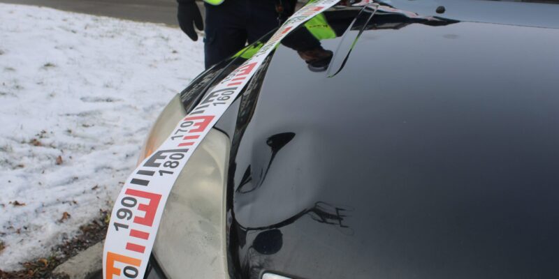 Uszkodzony samochód osobowy zabezpieczony policyjną taśmą na miejscu wypadku drogowego, w tle policjant w kamizelce odblaskowej i ośnieżone pobocze drogi.
