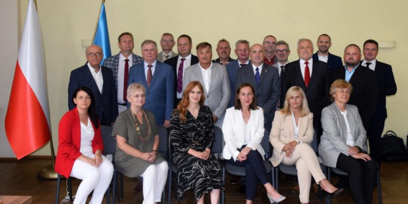 Radni Rady Miasta Oświęcim podczas wspólnego zdjęcia po sesji – grupa kilkunastu kobiet i mężczyzn w sali obrad, w tle flagi Polski i Unii Europejskiej.