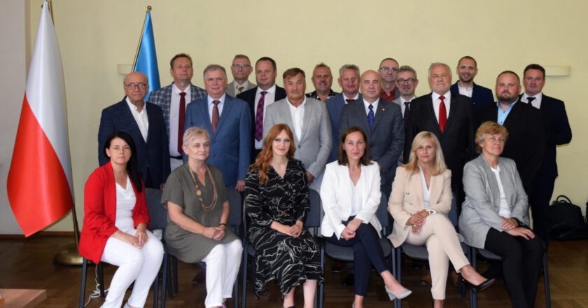 Radni Rady Miasta Oświęcim podczas wspólnego zdjęcia po sesji – grupa kilkunastu kobiet i mężczyzn w sali obrad, w tle flagi Polski i Unii Europejskiej.