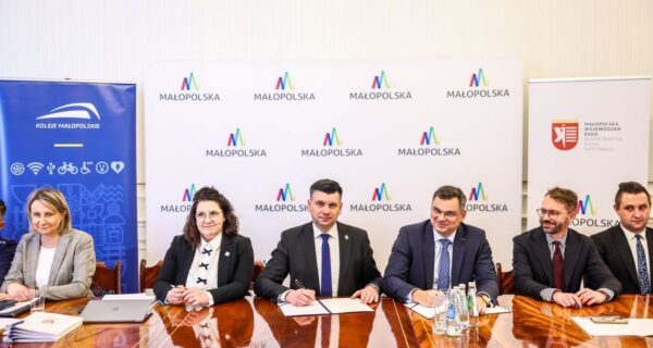 Podpisanie porozumienia o współpracy na rzecz bezpieczeństwa ruchu drogowego w Małopolsce – przedstawiciele władz województwa, Kolei Małopolskich i Wojewódzkiej Rady BRD siedzą przy stole konferencyjnym z logotypami Małopolski w tle.