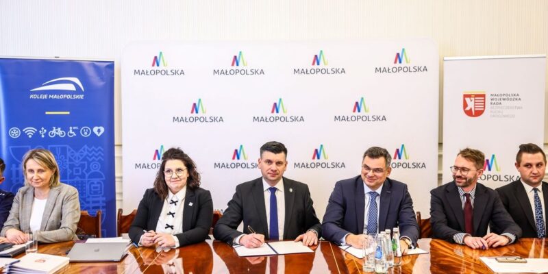 Podpisanie porozumienia o współpracy na rzecz bezpieczeństwa ruchu drogowego w Małopolsce – przedstawiciele władz województwa, Kolei Małopolskich i Wojewódzkiej Rady BRD siedzą przy stole konferencyjnym z logotypami Małopolski w tle.
