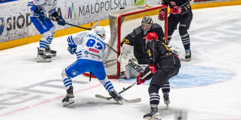 Akcja pod bramką Comarch Cracovii podczas meczu hokejowego z Unią Oświęcim. Ołeksandr Peresunko w biało-niebieskich barwach oddaje strzał z bliska, naciskany przez obrońców Cracovii, przed interweniującym bramkarzem krakowskiego zespołu. Dynamiczna sytuacja z derbów Małopolski w TAURON Hokej Lidze.