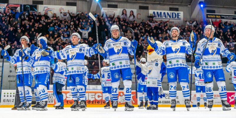 Hokeiści Unii Oświęcim dziękują kibicom na lodowisku po wysokim zwycięstwie, unosząc kije i bijąc brawo w stronę trybun wypełnionych fanami.