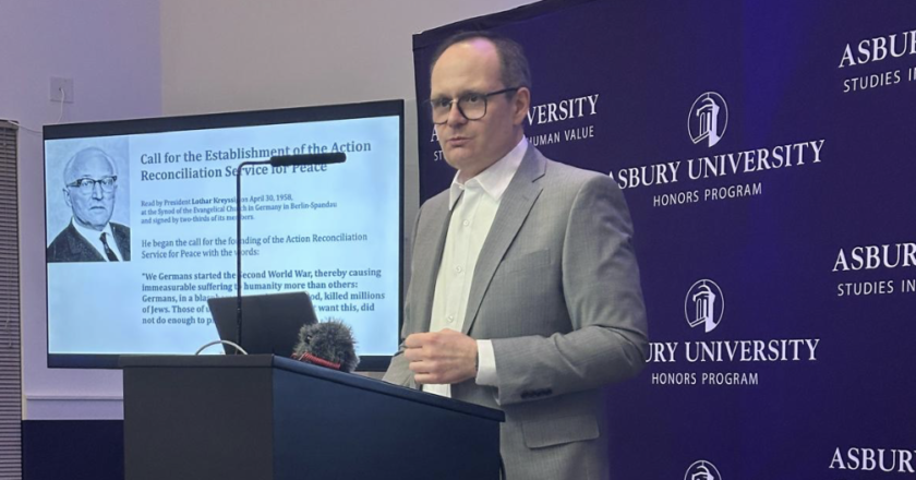 David Kennedy podczas wykładu na Asbury University w stanie Kentucky. Edukator Międzynarodowego Domu Spotkań Młodzieży w Oświęcimiu stoi przy mównicy, a w tle widoczne jest logo Asbury University oraz slajd prezentacji dotyczącej pojednania i edukacji historycznej.