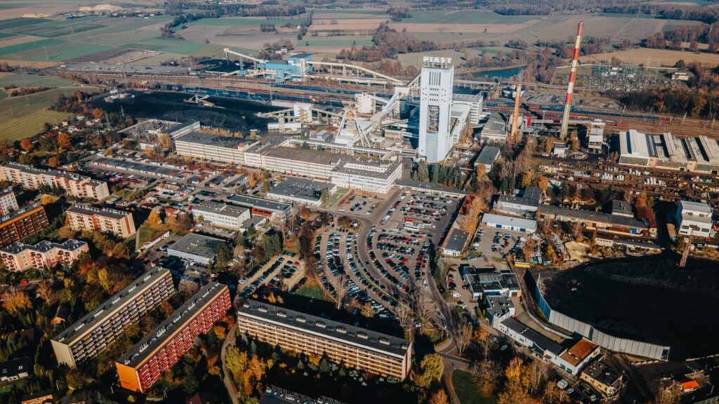 Widok z lotu ptaka na teren kopalni KWK Piast w Bieruniu – zabudowania przemysłowe, szyby, infrastruktura kolejowa oraz okoliczne osiedla mieszkaniowe i parkingi, w tle pola i tereny zielone.