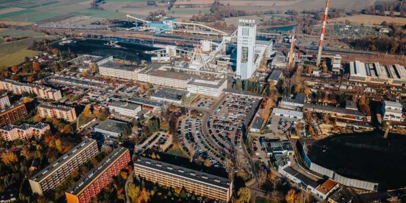 Widok z lotu ptaka na teren kopalni KWK Piast w Bieruniu – zabudowania przemysłowe, szyby, infrastruktura kolejowa oraz okoliczne osiedla mieszkaniowe i parkingi, w tle pola i tereny zielone.