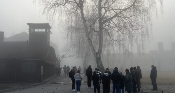 Grupa odwiedzających stoi przed bramą byłego obozu Auschwitz I w Oświęcimiu we mgle; widoczna wieża wartownicza i napis „Arbeit macht frei”, zimowa aura i ograniczona widoczność.