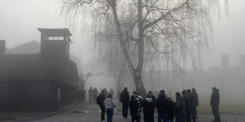 Grupa odwiedzających stoi przed bramą byłego obozu Auschwitz I w Oświęcimiu we mgle; widoczna wieża wartownicza i napis „Arbeit macht frei”, zimowa aura i ograniczona widoczność.