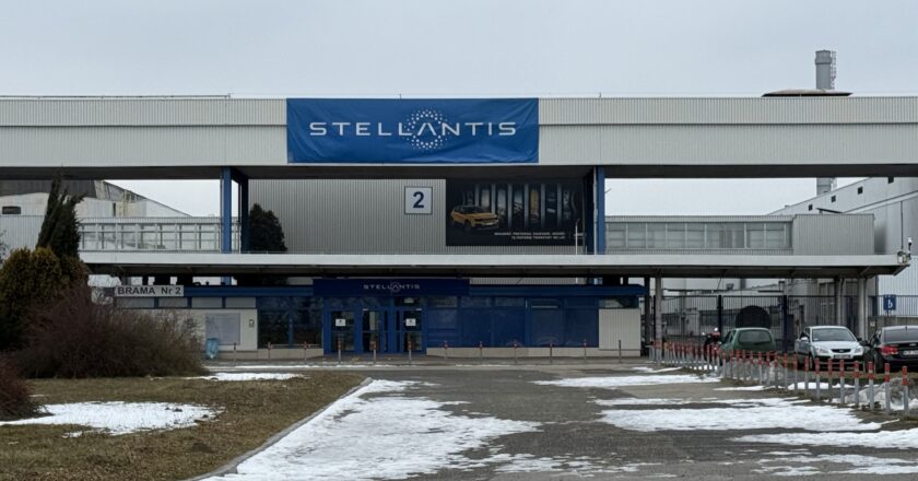 Brama nr 2 fabryki Stellantis w Tychach – na budynku widoczny duży niebieski baner z logo firmy. Przed wjazdem pusty plac i droga z resztkami śniegu, po prawej zaparkowane samochody.