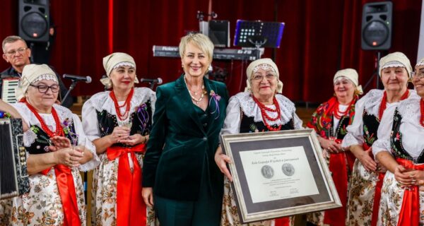 Uroczystość jubileuszowa Koła Gospodyń Wiejskich w Jawiszowicach. Na zdjęciu grupa kobiet w tradycyjnych strojach ludowych stoi na scenie z czerwonymi kotarami. W centrum widoczna jest Iwona Gibas w zielonym garniturze, a obok niej jedna z członkiń KGW trzyma w rękach duży, oprawiony dyplom dotyczący przyznania Srebrnego Medalu Polonia Minor. W tle widać sprzęt muzyczny i instrumenty.