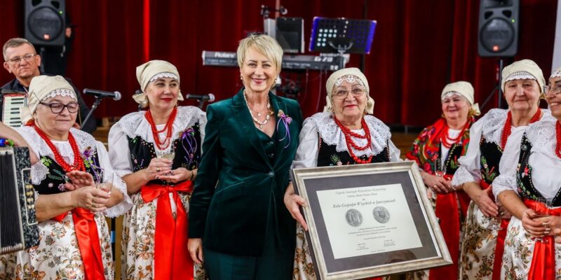 Uroczystość jubileuszowa Koła Gospodyń Wiejskich w Jawiszowicach. Na zdjęciu grupa kobiet w tradycyjnych strojach ludowych stoi na scenie z czerwonymi kotarami. W centrum widoczna jest Iwona Gibas w zielonym garniturze, a obok niej jedna z członkiń KGW trzyma w rękach duży, oprawiony dyplom dotyczący przyznania Srebrnego Medalu Polonia Minor. W tle widać sprzęt muzyczny i instrumenty.