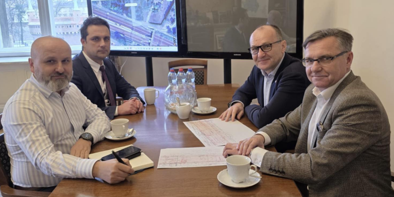 Spotkanie robocze w urzędzie – czterech mężczyzn siedzi przy stole konferencyjnym z dokumentami i kawą. W tle ekrany z mapą/schematem inwestycji drogowej.