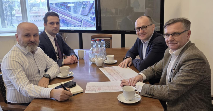 Spotkanie robocze w urzędzie – czterech mężczyzn siedzi przy stole konferencyjnym z dokumentami i kawą. W tle ekrany z mapą/schematem inwestycji drogowej.