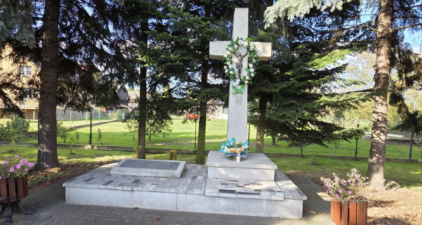 Pomnik w formie dużego kamiennego krzyża z figurą Jezusa, ozdobiony wieńcem i kwiatami, stojący na granitowym cokole wśród drzew.