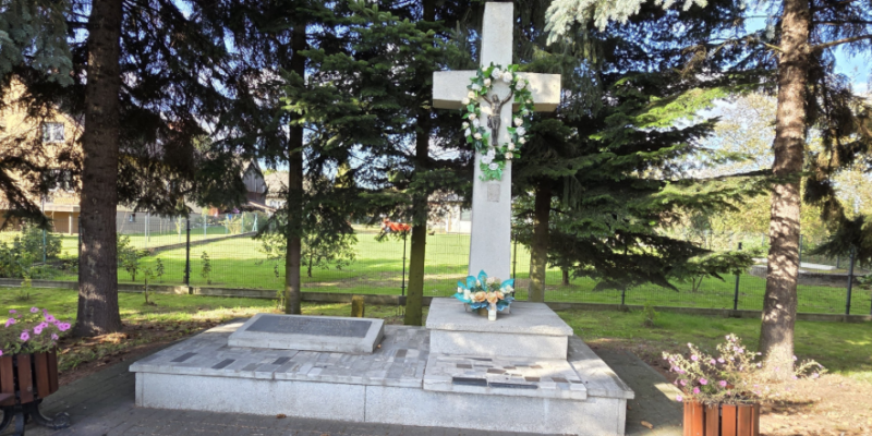 Pomnik w formie dużego kamiennego krzyża z figurą Jezusa, ozdobiony wieńcem i kwiatami, stojący na granitowym cokole wśród drzew.
