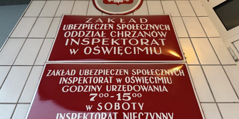 Tablica na elewacji budynku Zakładu Ubezpieczeń Społecznych, Inspektorat w Oświęcimiu (Oddział Chrzanów). Na białych kafelkach zamontowane dwie bordowe tablice z białym napisem oraz godło Polski. Dolna tablica informuje o godzinach urzędowania: 7:00–15:00, w soboty inspektorat nieczynny.